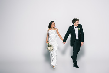 full length view of happy, elegant interracial newlyweds holding hands while walking and looking away on white background