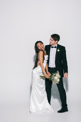 full length view of elegant bridegroom hugging happy african american bride holding wedding bouquet on white background