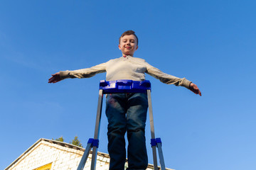 A teenage boy stands on a high ladder stepladder and raised his hands to fly into the sky.