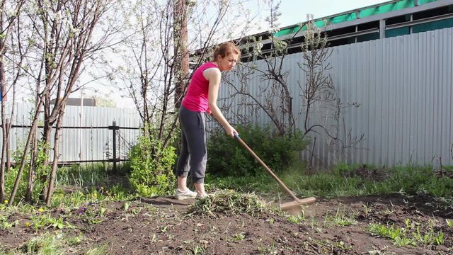 A young woman on the plot of land removes the grass with a rake.