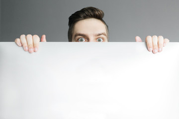 Young man peeping from behind white wall on grey background