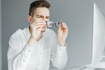 Young man in white shirt looking through glasses. Worker at the desktop with computer on grey background
