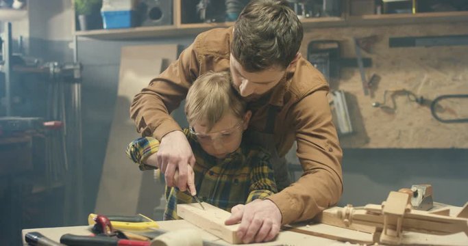 Caucasian handsome father carpenter teaching small cute son to carve wood in workshop. Woodman showing little boy how to carving on timber. Handmade carpentry work.