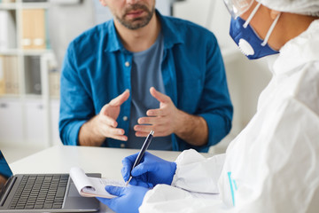 Close-up of doctor in protective clothing sitting at the table and writing prescription with...