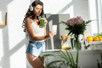 Selective focus of woman in headphones taking selfie with smartphone while holding smoothie in kitchen
