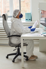 Fototapeta premium Female doctor in protective workwear sitting at the table working on computer and examining documents at office