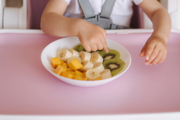 Obraz na płótnie Canvas Close up of baby girl take exotic fruits from plate in high chair. Sliced mango, kiwi and banana