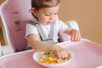 Beautiful little baby girl first time eating exotic fruits in high chair. Cute baby girl taste delitious fruist, mango, kiwi, and babana