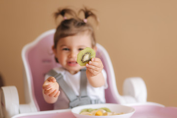 Obraz na płótnie Canvas Beautiful little baby girl first time eating exotic fruits in high chair. Cute baby girl taste delitious fruist, mango, kiwi, and babana