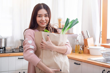 Young Asian woman in the kitchen and holding grocery shopping bag, looking at camera, carried brown paper bags in the kitchen