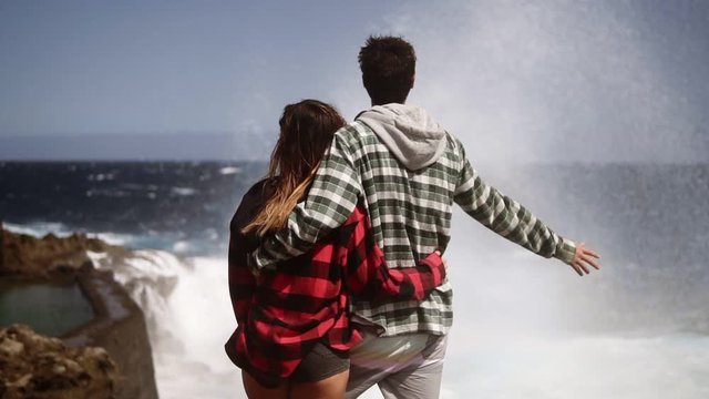 Young love couple consisting of man and woman in plaid coats standing near stormy sea, ocean rocky coast, watching big foamy waves coming and breaking on rocks. Rare view