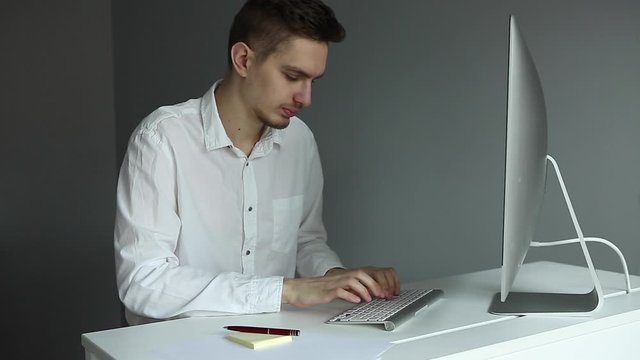 Young man in white shirt working at the desktop with a computer in office. Worker on grey background