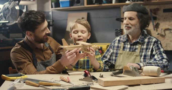 Portrait of three male generations smiling to camera and showing handmade wooden plane. Grandfather, father and son of cheerful carpenters in workshop. Old man with son and grandson. Family of joiners