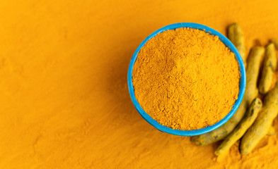 Turmeric powder in blue bowl with dry roots at orange background.