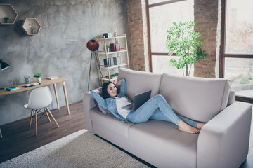 Nice attractive cheerful cheery slim fit thin girl lying on divan using laptop studying watching web tutorial spending free time in modern loft brick industrial house apartment