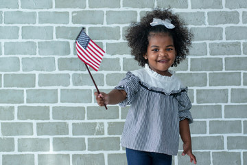 Dark-skinned little girl smiling and waving American flag and looking at camera, holding flag at...