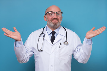 Mature male doctor shrugging lifting shoulders having no idea how to cure on blue background.
