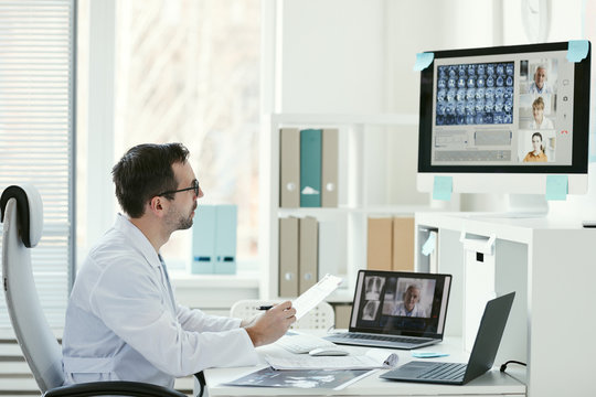 Mature Male Doctor Looking At The Computer Monitor And Talking To His Colleagues Online At Office