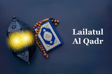 LAILATUL QADR text with Holy Al Quran with written arabic calligraphy meaning of Al Quran, lantern lamp and rosary beads or tasbih on black background.