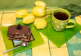 On the table is a cup of tea and a plate with a slice of cake and a spoon. Nearby are yellow tulips.