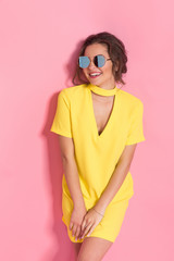 Beautiful girl wearing yellow dress and sunglasses posing on pink background in studio.