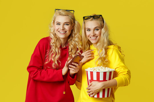 Shocked young models women in 3d glasses eating popcorn, looks Movie at the cinema. Isolated on yellow background