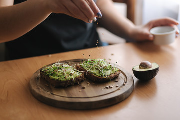 Woman making vegan sandwich using rye toasted bread with guacamole and micro greens. Woman sprinkle with sesame seeds on sandwich