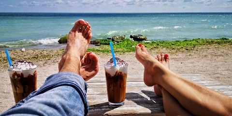 Young barefoot couple relaxing and drinking ice coffee cocktails at summer beach cafe bar on a bright sunny day
