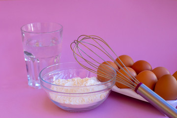 Products for the preparation of dough. Eggs, flour, water, whisk. Pink background