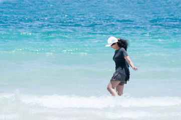 Cute Asian girl wearing a black t-shirt and white hat standing on the sunny tropical beach relaxing in water , Beautiful young woman feeling funny on the sea.