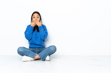 Young mixed race woman sitting on the floor isolated on white background covering mouth with hands