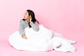 Young mixed race woman wearing pijama sitting on the floor listening to something by putting hand on the ear