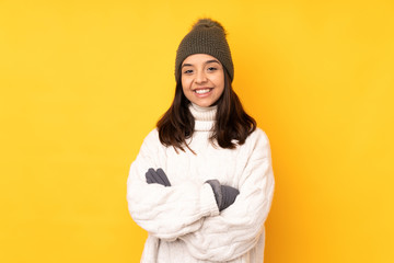 Young woman with winter hat over isolated yellow background keeping the arms crossed in frontal...