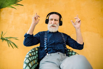 Studio shot of cool good-looking elderly man with well-groomed beard, sitting on the chair and enjoying his favourite music in earphones. Isolated on yellow background