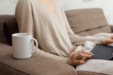 A white mug stands on the arm of a brown sofa against the blurry background of a girl's hands are typing on a laptop. Concept of distance work.