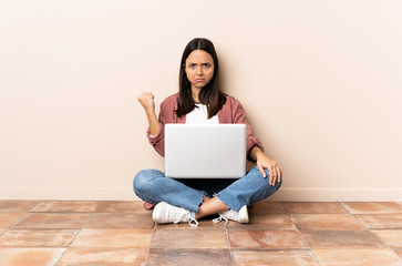 Young mixed race woman with a laptop sitting on the floor with unhappy expression