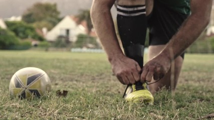Closeup of a rugby player tying shoes on field with a rugby ball on the side. Rugby player adjusting his shoe laces on ground.
 - Powered by Adobe