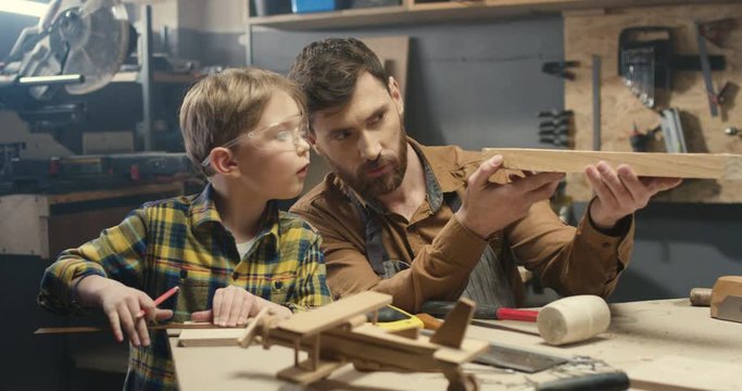 Cheerful handsome young Caucasian carpenter working at desk in workshop and small son playing next to him. Faher woodman proceeding wood at table with little boy helping.