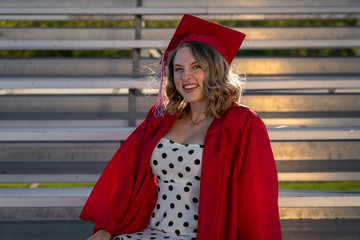 A cute girl in a polka dot dress and cap and gown sits on bleachers and smiles for a graduation portrait picture.