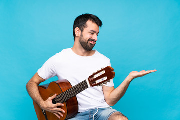 Young man with guitar over isolated blue background holding copyspace with doubts