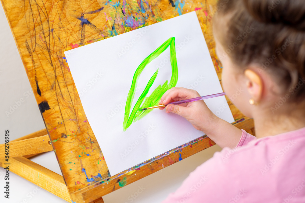 Wall mural Child draws green leaf by watercolors on white paper on an easel. Earth day and environmental protection concept.