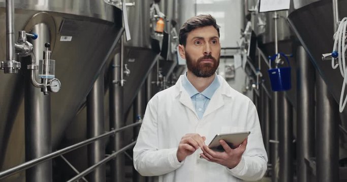Bearded man in white lab coat entering data on tablet at brewery. Attentive male worker touching and typing digital display while walking and looking on automatic beverage equipment