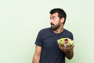 Young handsome man with salad over isolated green wall making doubts gesture looking side