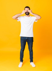Full-length shot of man with beard over isolated yellow background covering eyes by hands
