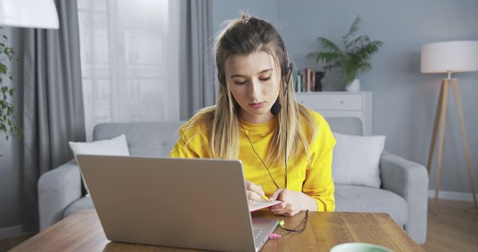 Female student attending online classes, making notes in a beautiful red notebook, using black headphones to communicate. Сoncept of online learning, online courses, remote individual lessons.
