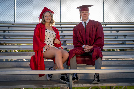 A Pretty Girl And Handsome Boy In Red And Maroon Caps And Gowns Sit Together On Bleachers To Celebrate And Commemorate Graduation.