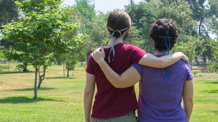 two Caucasian women viewed from behind with their arms around each other's shoulders at a lush green park on a sunny day with copy space