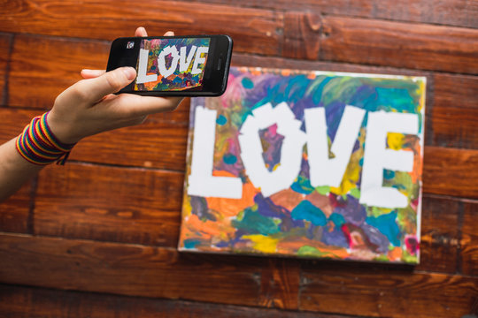 Homosexual woman photographs with her phone a watercolor that she has painted with the word Love.
