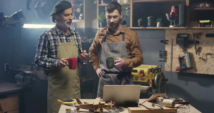 Old and young male carpenters watching video on laptop computer in workshop while smoking and sipping hot drinks. Senior father and son, handicraftsmen resting and drinking coffee or tea in workroom.