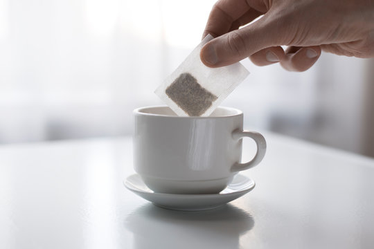 Close-up of a hand putting a tea bag into a white ceramic cup on a saucer. Morning beverage preparation and relaxation concept.